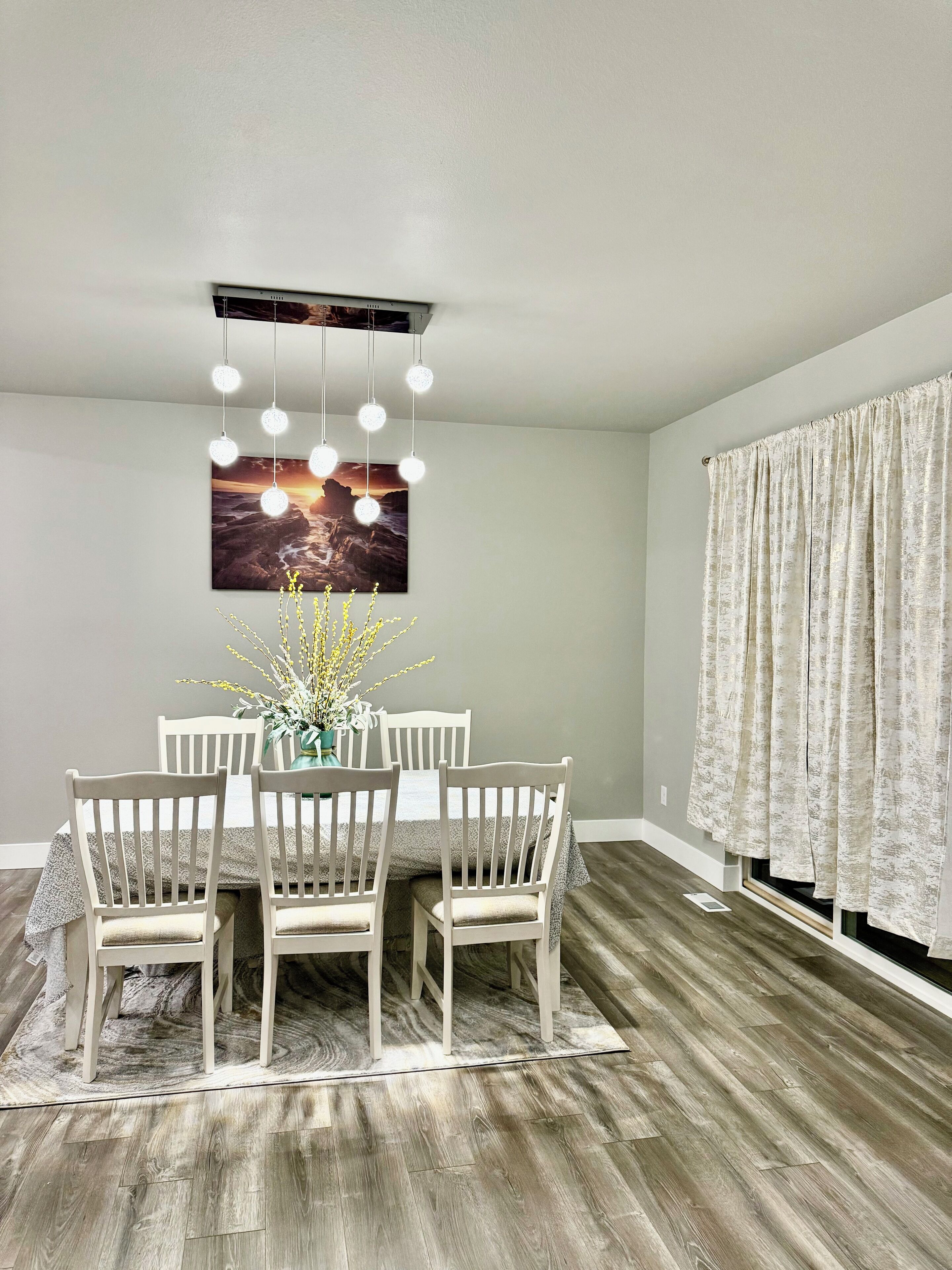 A dining area with a table set for four, a hanging light fixture, and a window with curtains.