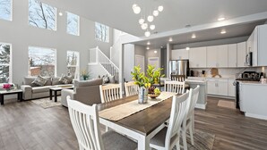 A spacious living area with a wooden dining table, white chairs, and a sofa with cushions. A vase with flowers is on the table. - LE'S SINGLE HOUSE WITH LAKE VIEW (Anchorage)