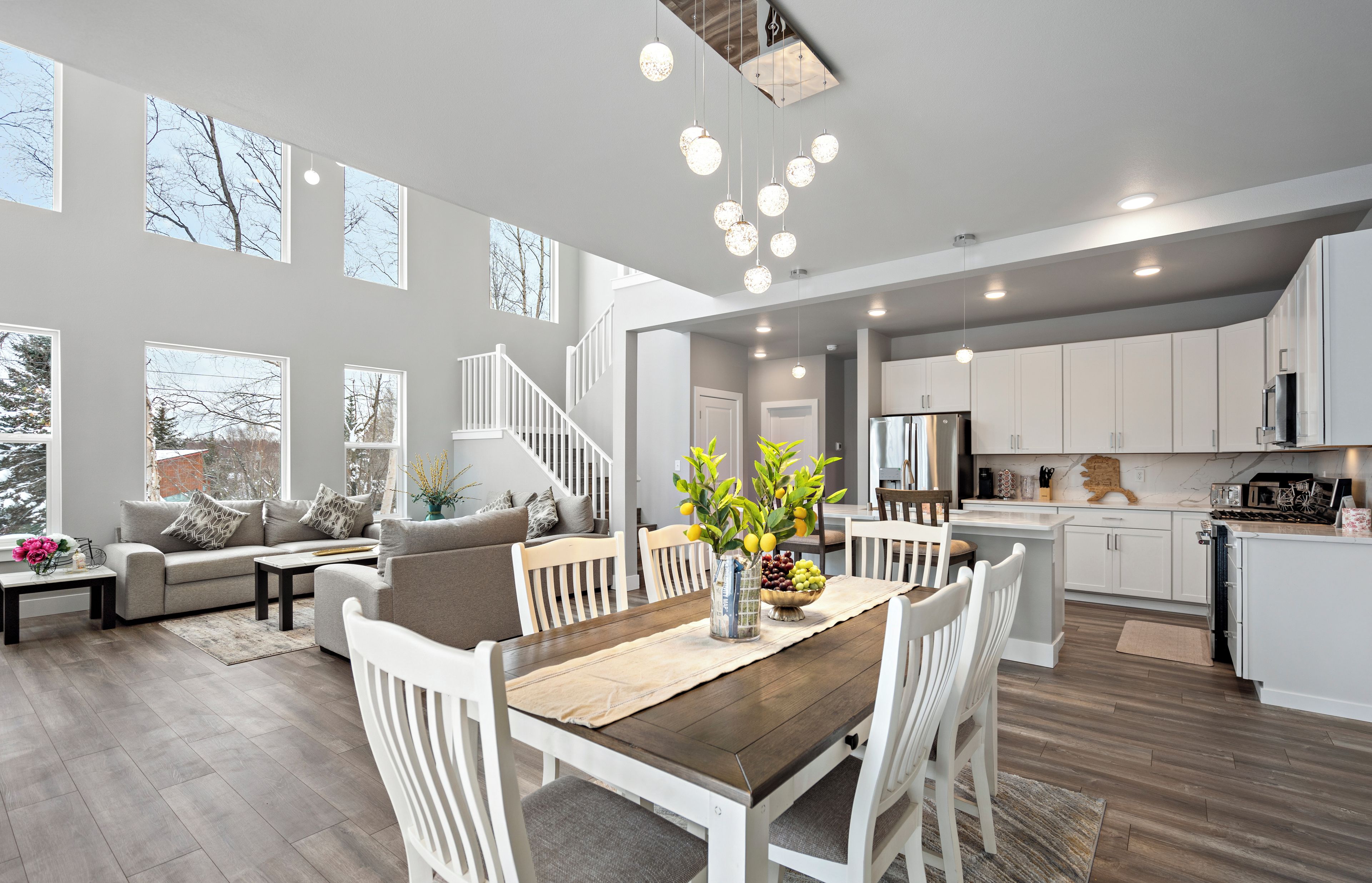 A spacious living area with a wooden dining table, white chairs, and a sofa with cushions. A vase with flowers is on the table.