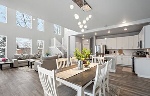 Een ruime woonkamer met een houten eettafel, witte stoelen en een bank met kussens. Op de tafel staat een vaas met bloemen.