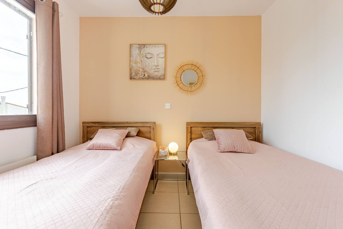 Una habitació d'hotel amb dos llits, una finestra amb cortines, una paret amb un quadre i un mirall, i una taula de nit amb una làmpada.