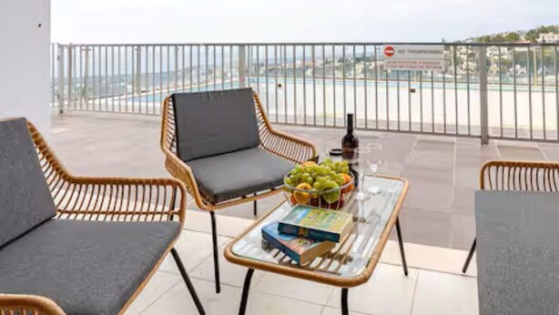 Varanda com cadeiras de vime, mesa de vidro com um livro e frutas, e vista para a praia e piscina.