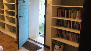 Een gang met een blauwe deur die uitkomt op een tuin, een witte boekenkast vol boeken, en een betegelde vloer.