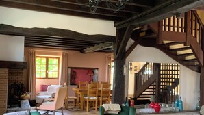 Een ruime woonkamer met hoge plafonds, zichtbare houten balken en een open haard. Er staan twee groene fauteuils, een houten eettafel met stoelen, en een trap die naar een hoger niveau leidt.