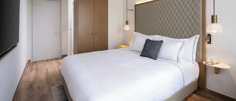 A modern hotel room with a large bed, a wardrobe, a bedside table, and a wall-mounted light fixture.
