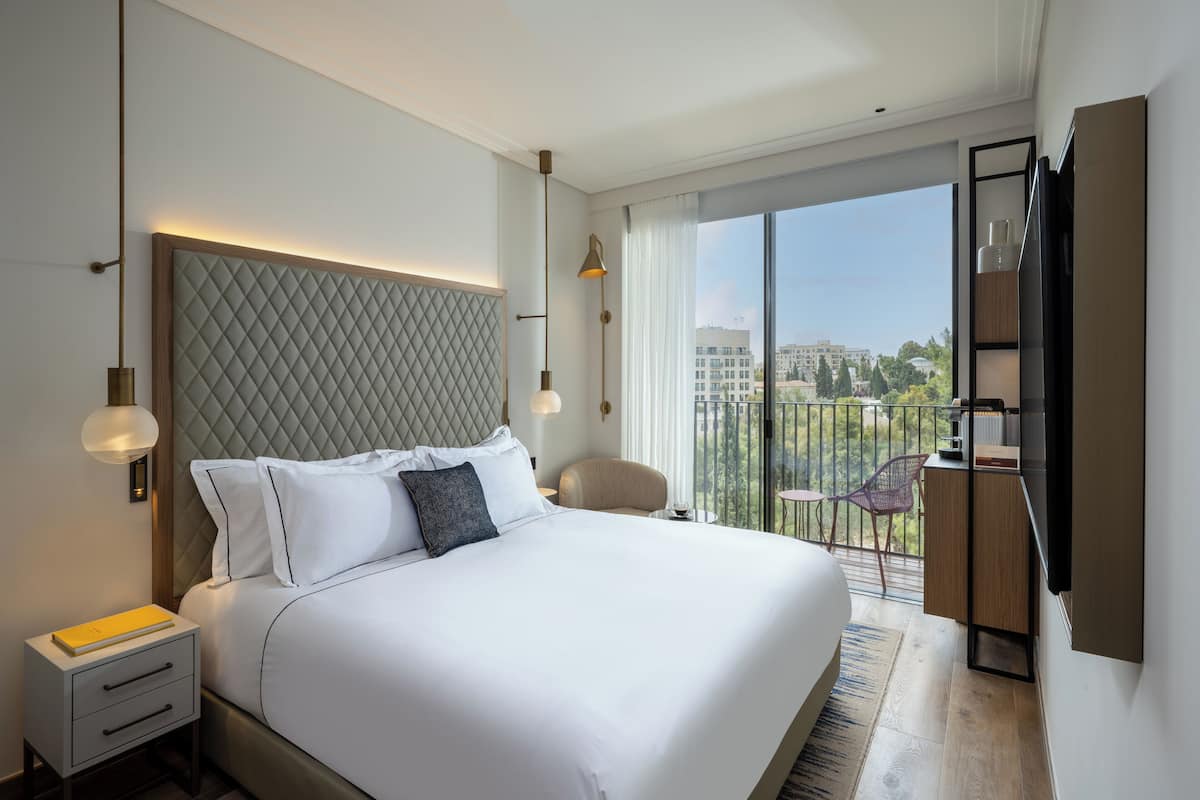 A modern hotel room with a large bed, a desk, a chair, and a balcony with a view of the city.