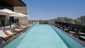 A rooftop pool with lounge chairs and umbrellas, surrounded by city buildings. - Hotel Nucha - FATTAL COLORS (Jerusalem)