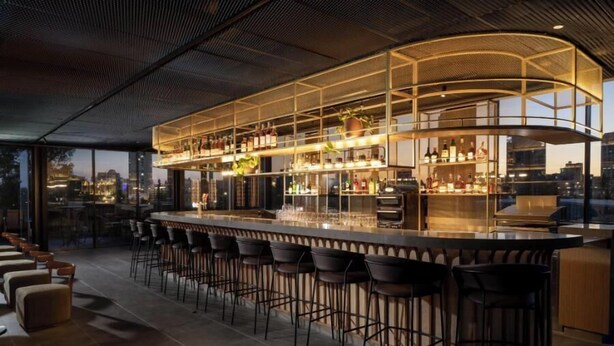 A modern bar area with a curved counter, multiple stools, and a city view. - Hotel Nucha - FATTAL COLORS (Jerusalem)