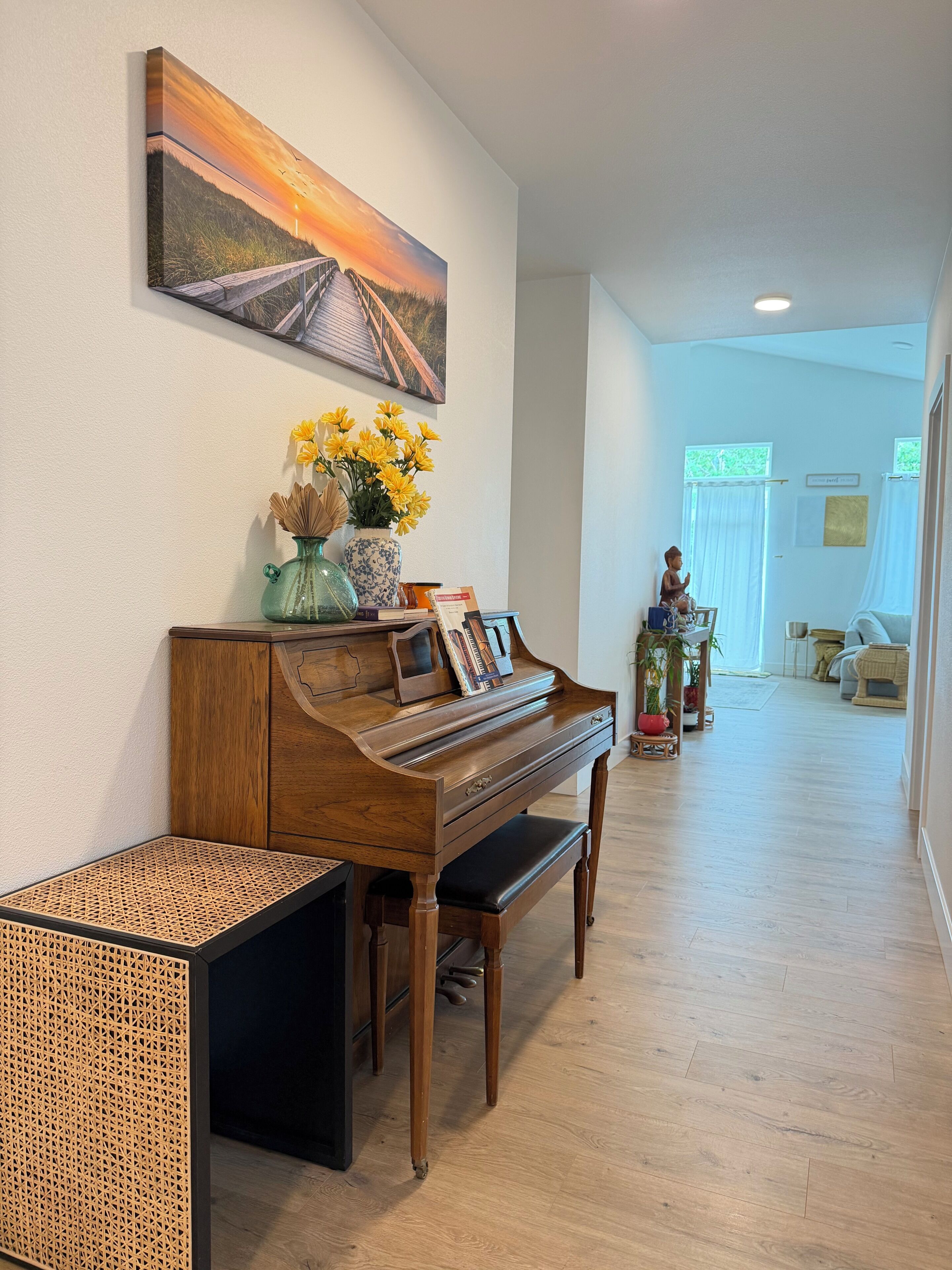 A piano with a decorative vase and books, a wooden bench, and a painting of a sunset on the wall.