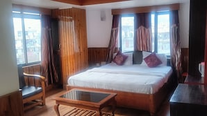 Ett hotellrum med en säng, en stol i trä, ett bord med glasskiva och utsikt över staden genom stora fönster.