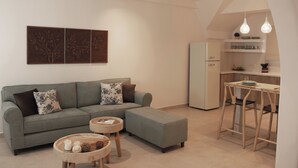 A modern living room with a grey sofa, ottoman, and round coffee table, a kitchen area with a white fridge and bar stools, and a dining table with hanging pendant lights. - Ostria, Pithos Apartments (Thira - Santorini)