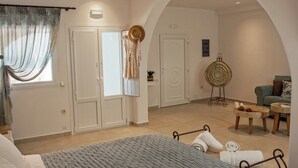 A bedroom with a bed, a wardrobe, a vanity, a round mirror, and a seating area with a sofa and side tables. - Ostria, Pithos Apartments (Thira - Santorini)