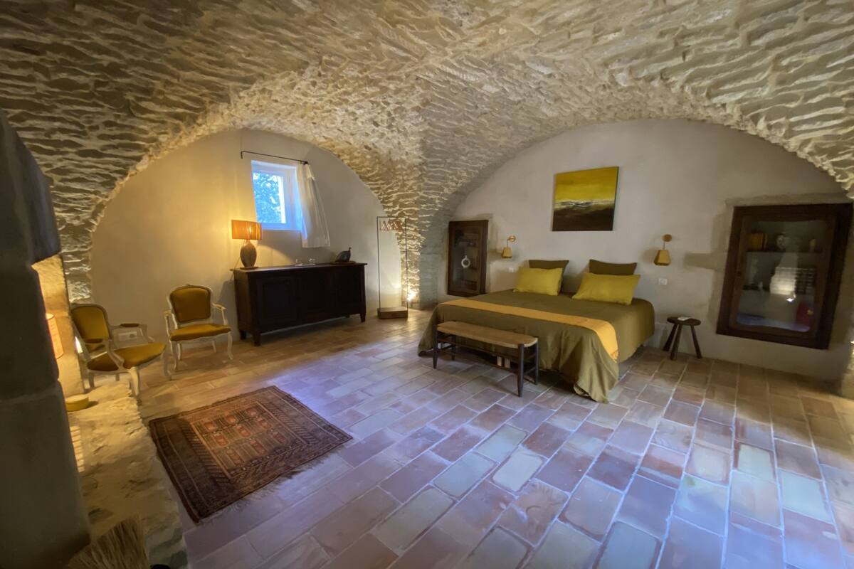 A cozy bedroom with a stone ceiling, a bed with yellow bedding, a bedside table, a mirror, and a window.