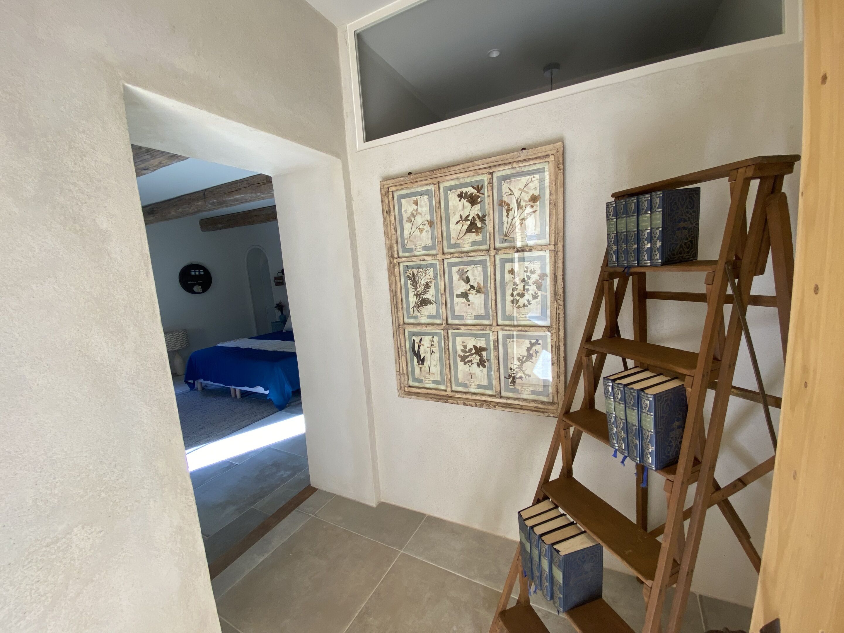 A room with a bed, a wooden ladder with books, and a wall with framed botanical prints.