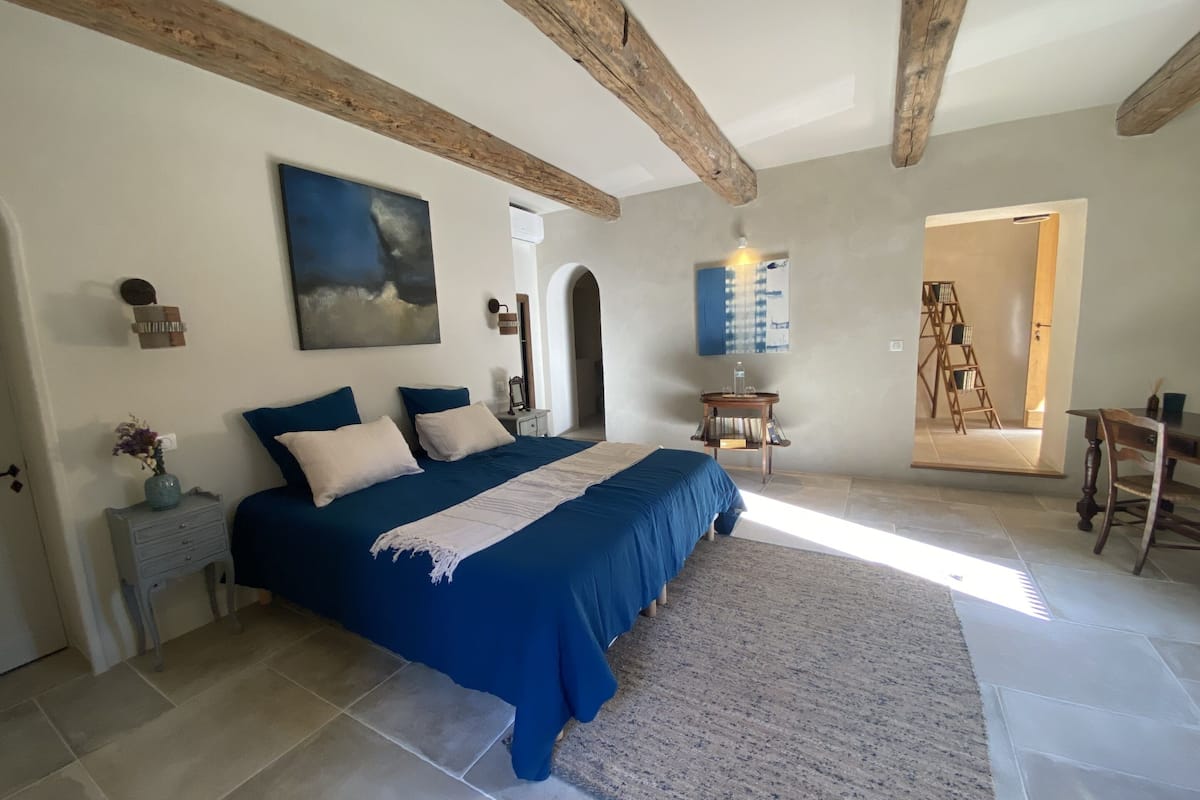 A bedroom with a large bed, a wooden ceiling, a painting, a small table with a chair, and a ladder in the doorway.