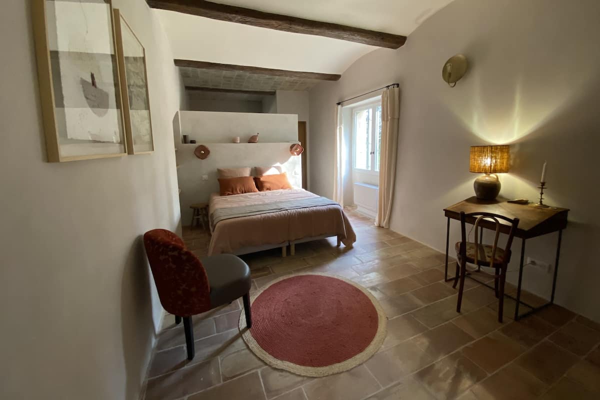 A bedroom with a bed, a desk, a chair, a red rug, and a window with curtains.