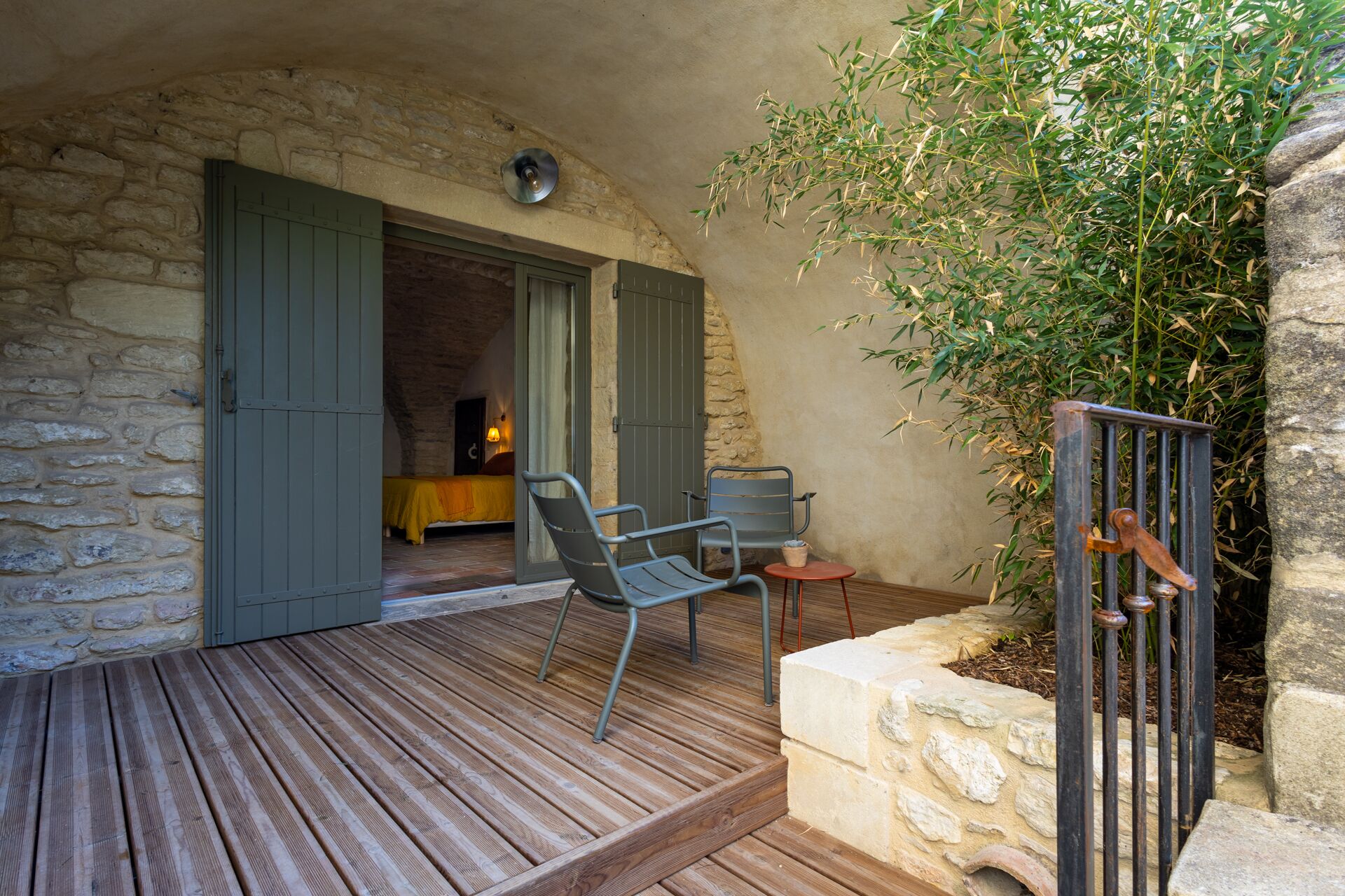 A wooden deck with metal chairs, a stone wall, and a door leading to a room with a bed and a table.