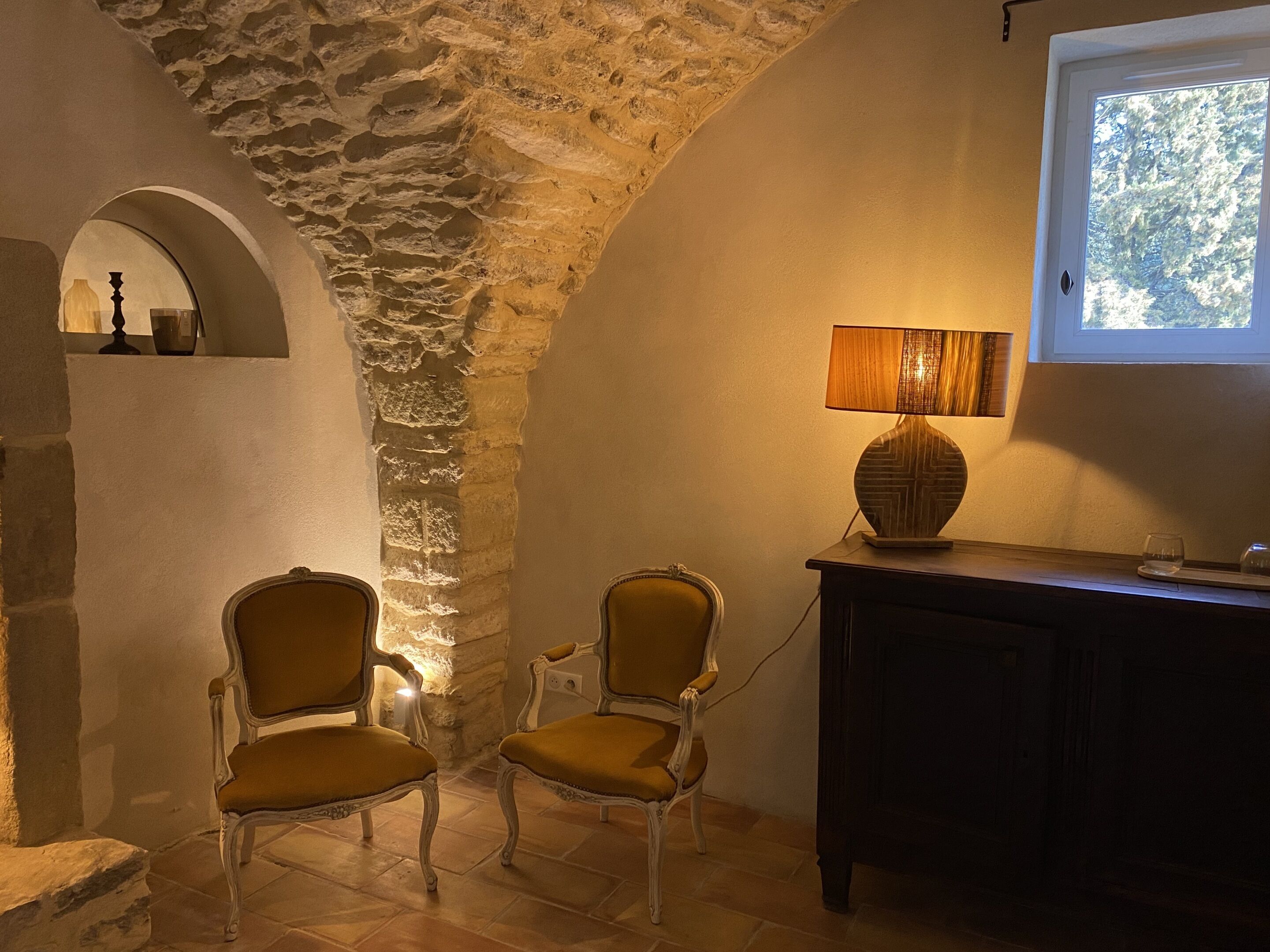 A room with a stone wall, two chairs, a lamp, and a window with a view of trees.