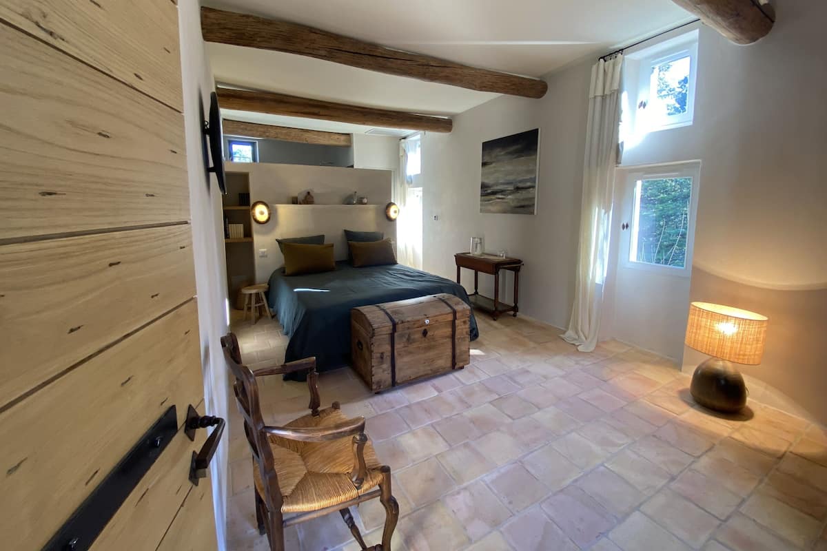 A bedroom with a bed, wooden headboard, bedside tables, a chair, a wooden chest, and a window with curtains.
