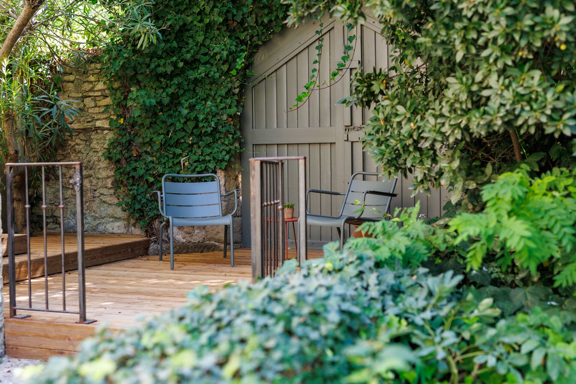 A wooden deck with two metal chairs and a small table, surrounded by lush greenery and a stone wall.
