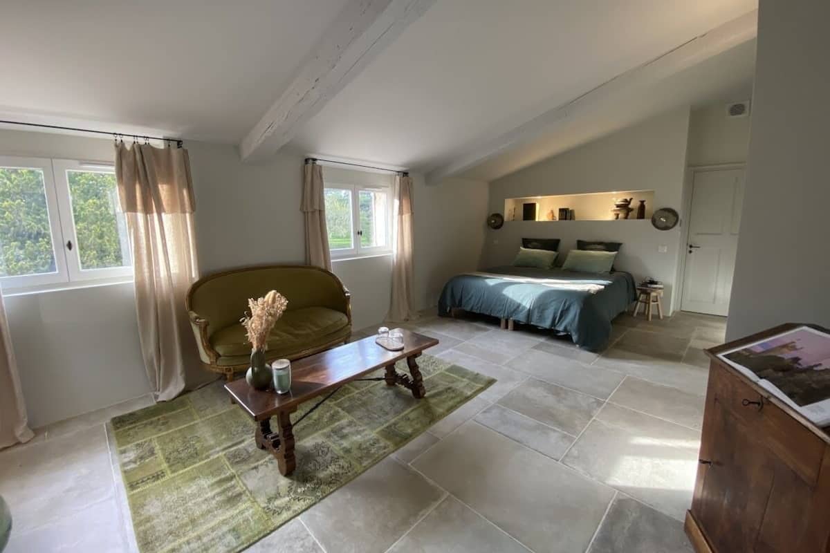 A spacious room with a large bed, a sofa, and a wooden cabinet. There is a window with curtains, a small table with a vase, and a rug on the floor.