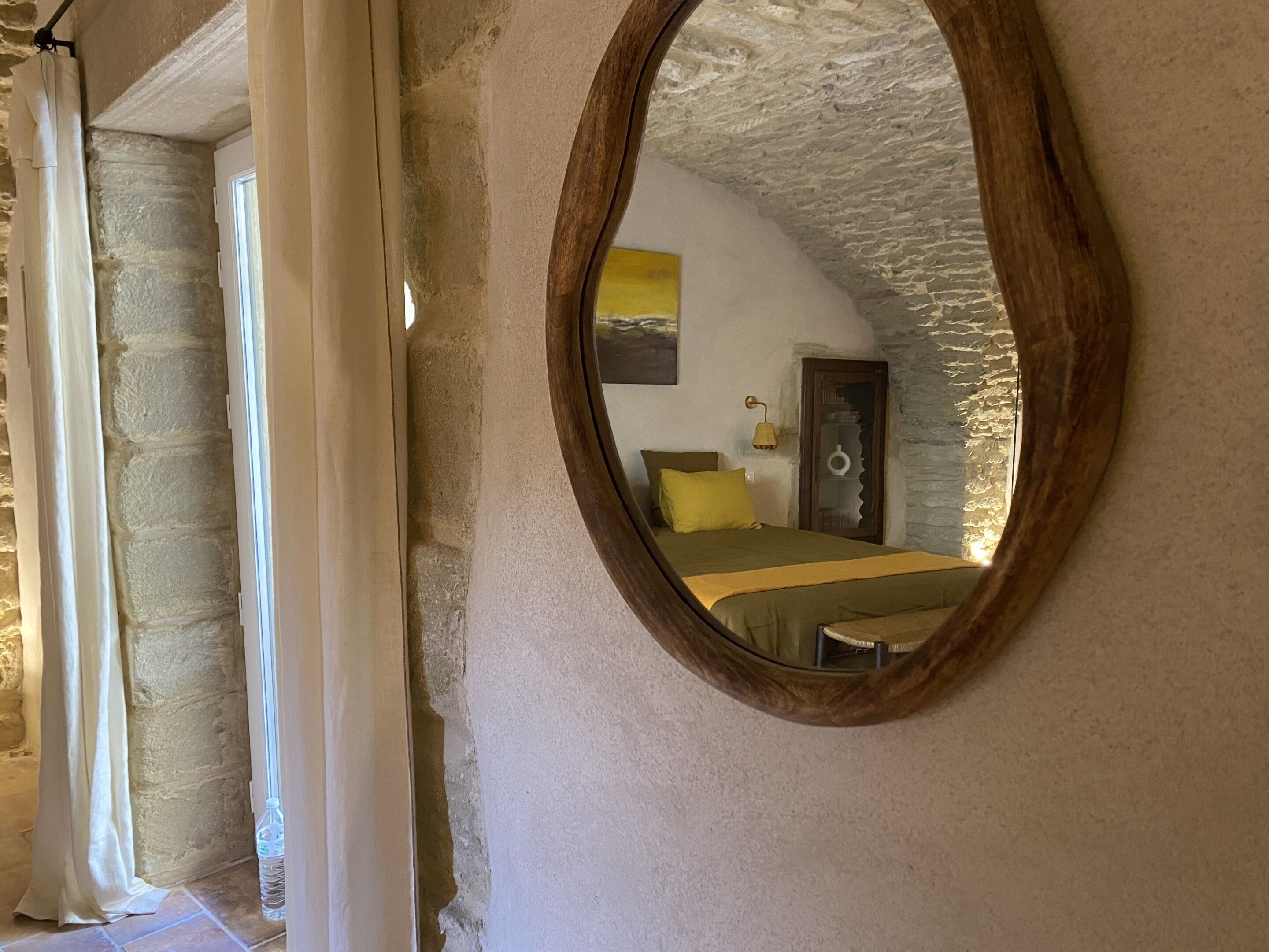 A bedroom with a stone wall, a bed with a green and yellow bedspread, and a wooden-framed mirror reflecting the room.