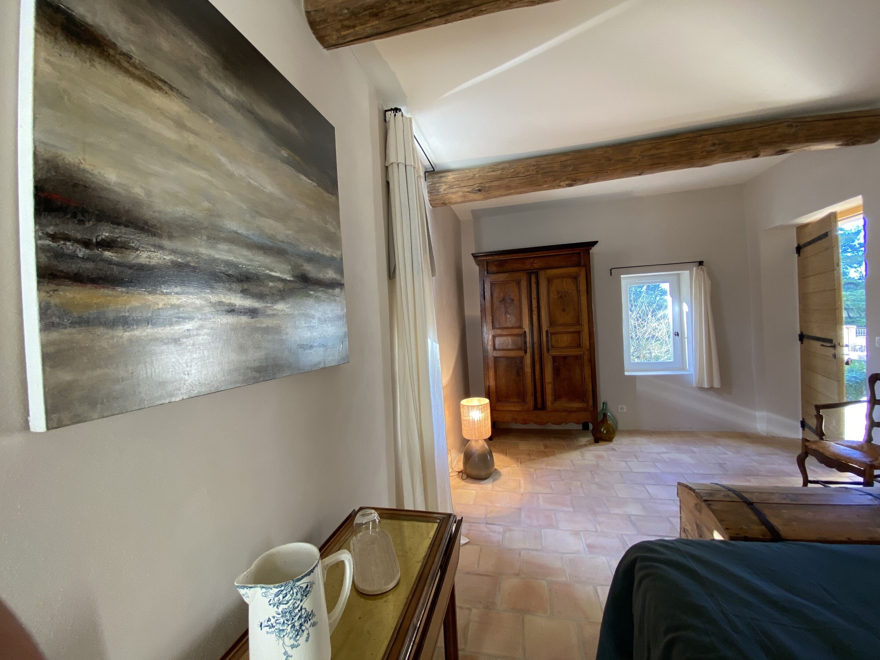 Una stanza con un grande quadro astratto, un armadio in legno, un tavolo con una brocca, una lampada e vista sull'esterno attraverso una finestra.