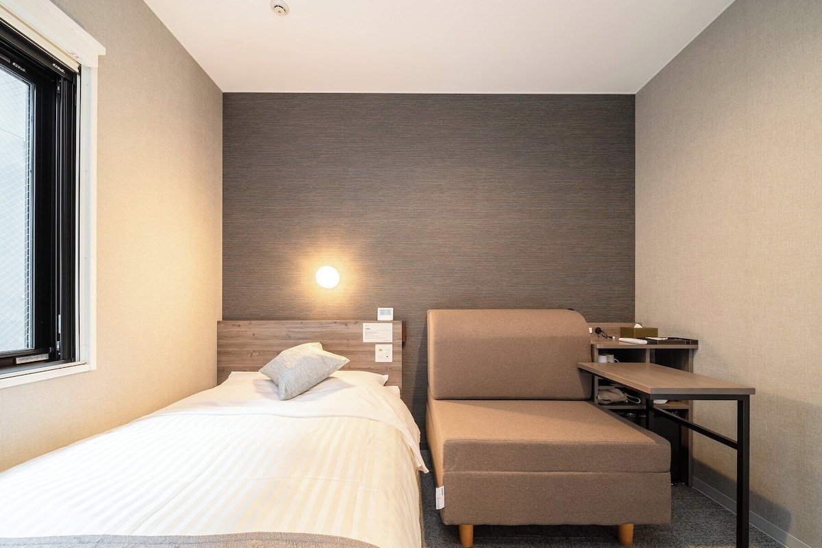 Une chambre d’hôtel moderne comprenant un lit, un coin salon avec une chaise et une table, et un luminaire fixé au mur.