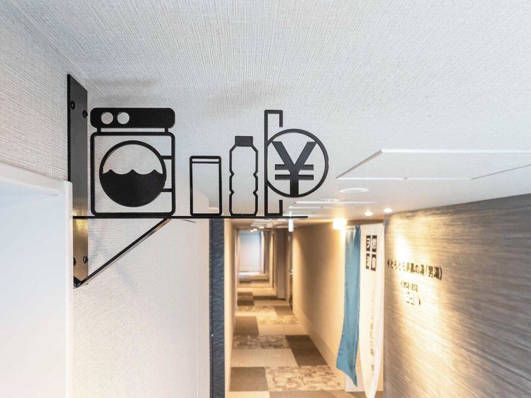 A hallway with a modern design, featuring a wall-mounted sign with icons representing amenities such as a washing machine, a bottle, and a yen symbol.