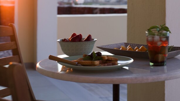 A table with a bowl of strawberries, a plate with bread, and a glass of iced drink, set against a backdrop of a flag and a sunset view. - Maistros, Pithos Apartments (Thira - Santorini)