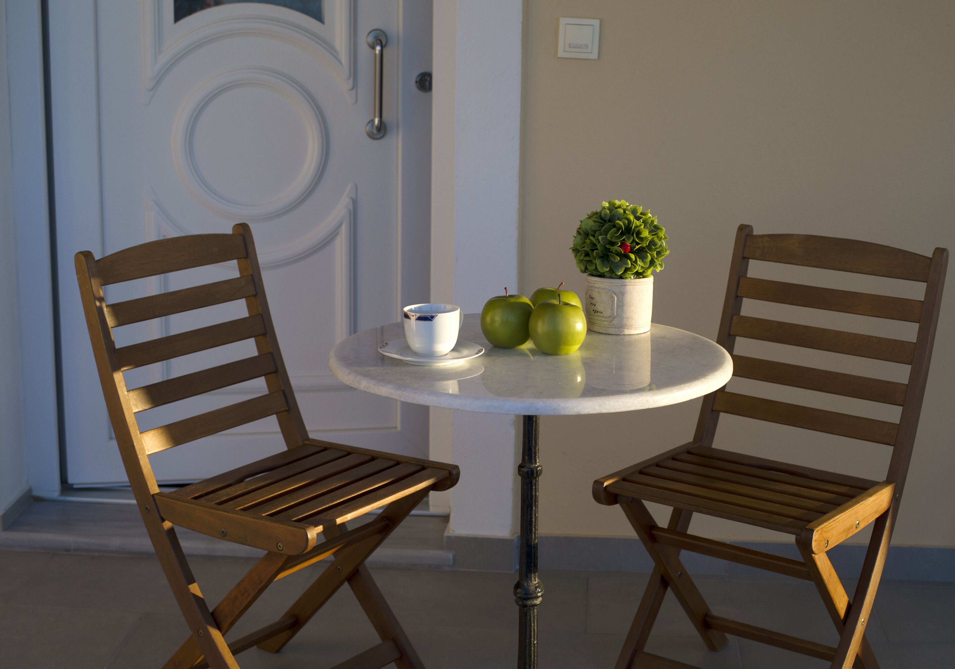 A round table with two wooden chairs, a white mug, and three green apples.