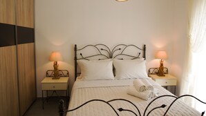 A neatly made bed with white linens and two bedside lamps. There is a wooden wardrobe to the left and a window with curtains to the right. - Maistros, Pithos Apartments (Thira - Santorini)