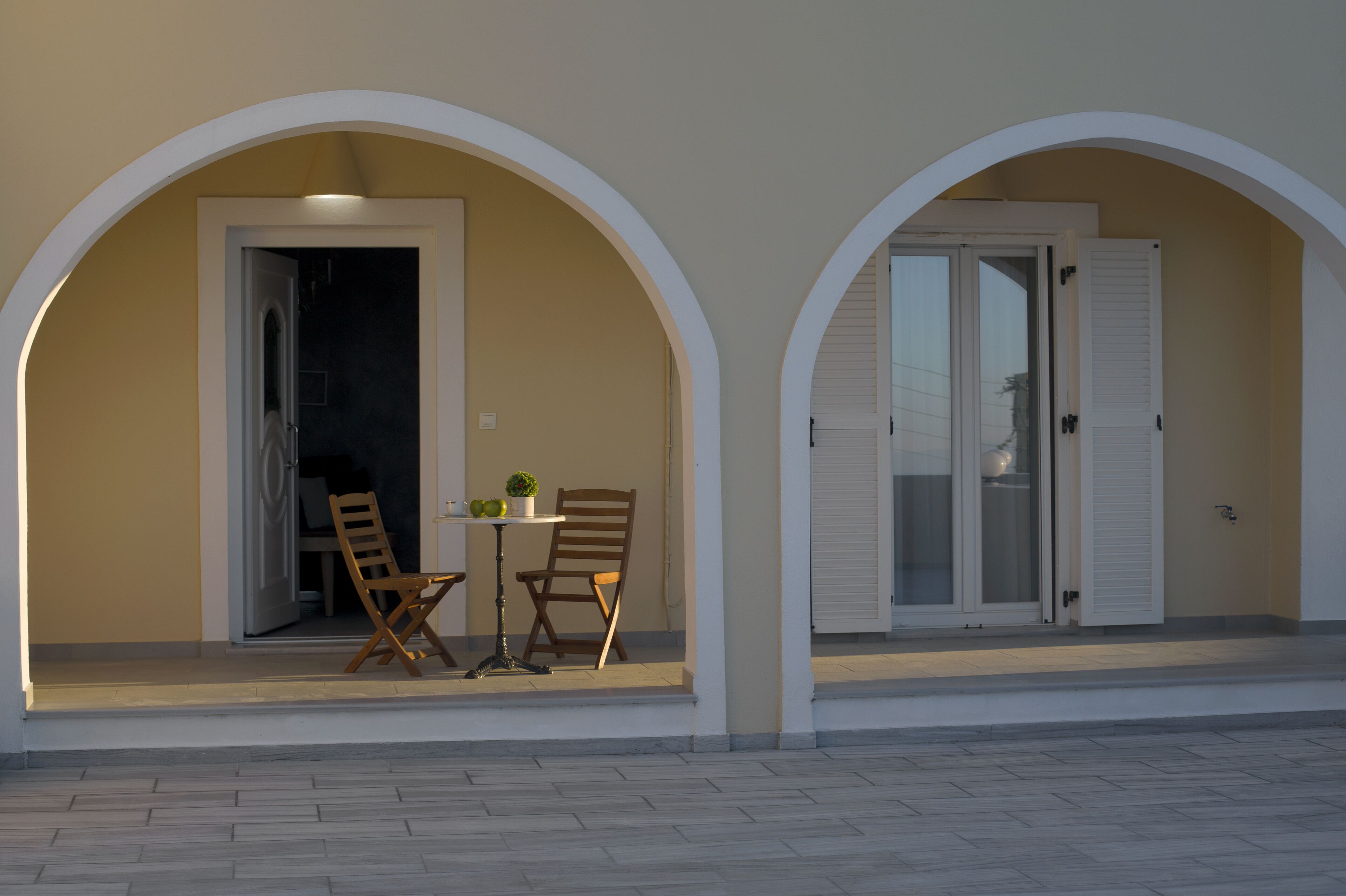 A covered outdoor seating area with two wooden chairs and a small table, connected to a building by an arched doorway.