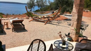 A hammock under a wooden pergola, a wooden lounge chair, and a table with chairs in a garden setting with a view of the sea.