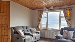 A living room with a grey sofa, a smaller armchair, wooden flooring, a wooden ceiling, and a view of the sea through a window. - Cottage 467 - Kilkerrin - sleeps 8 guests  in 4 bedrooms (Kilkerrin)
