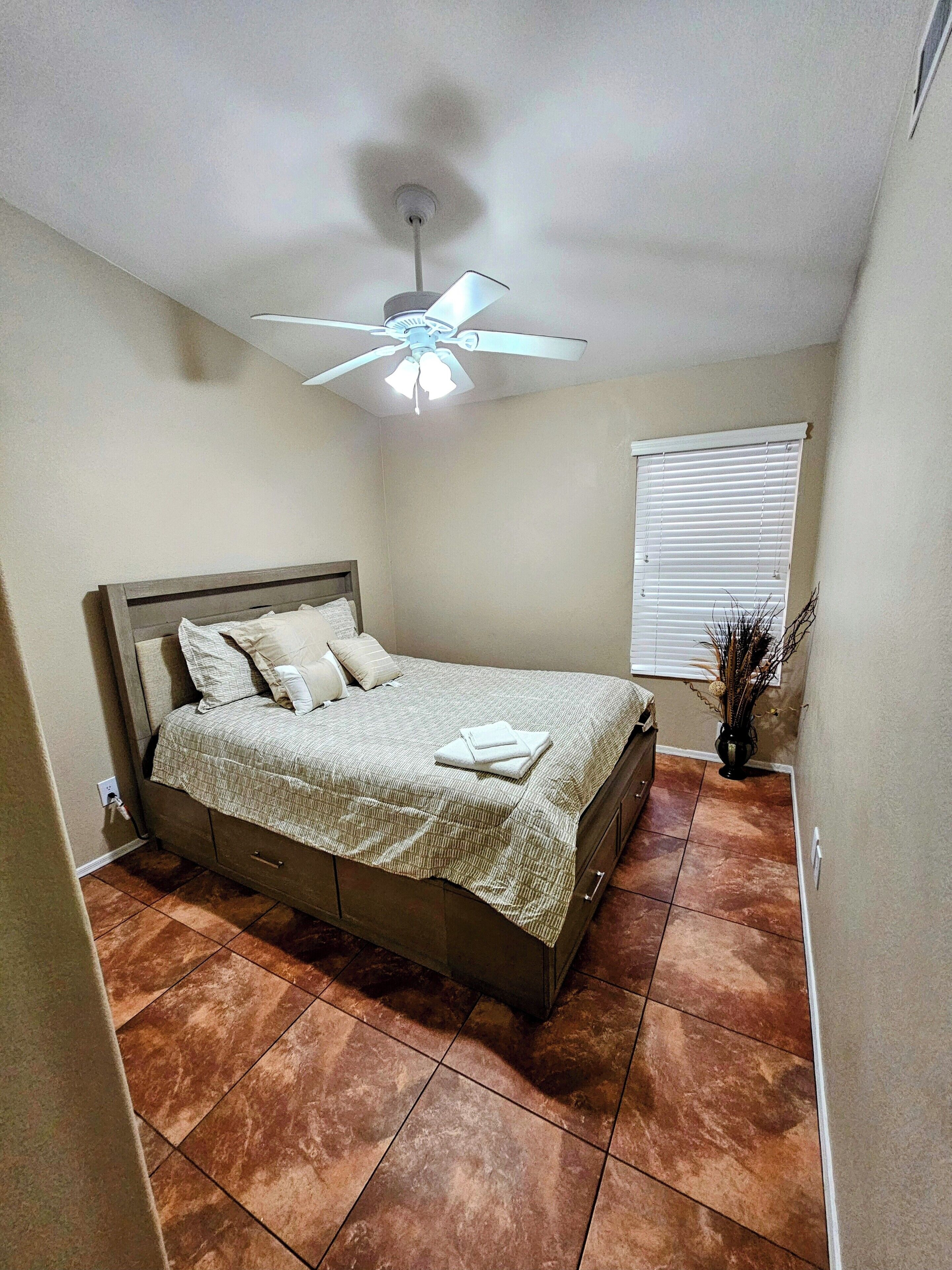 A bedroom with a bed, a ceiling fan, a window with blinds, and a vase with plants.