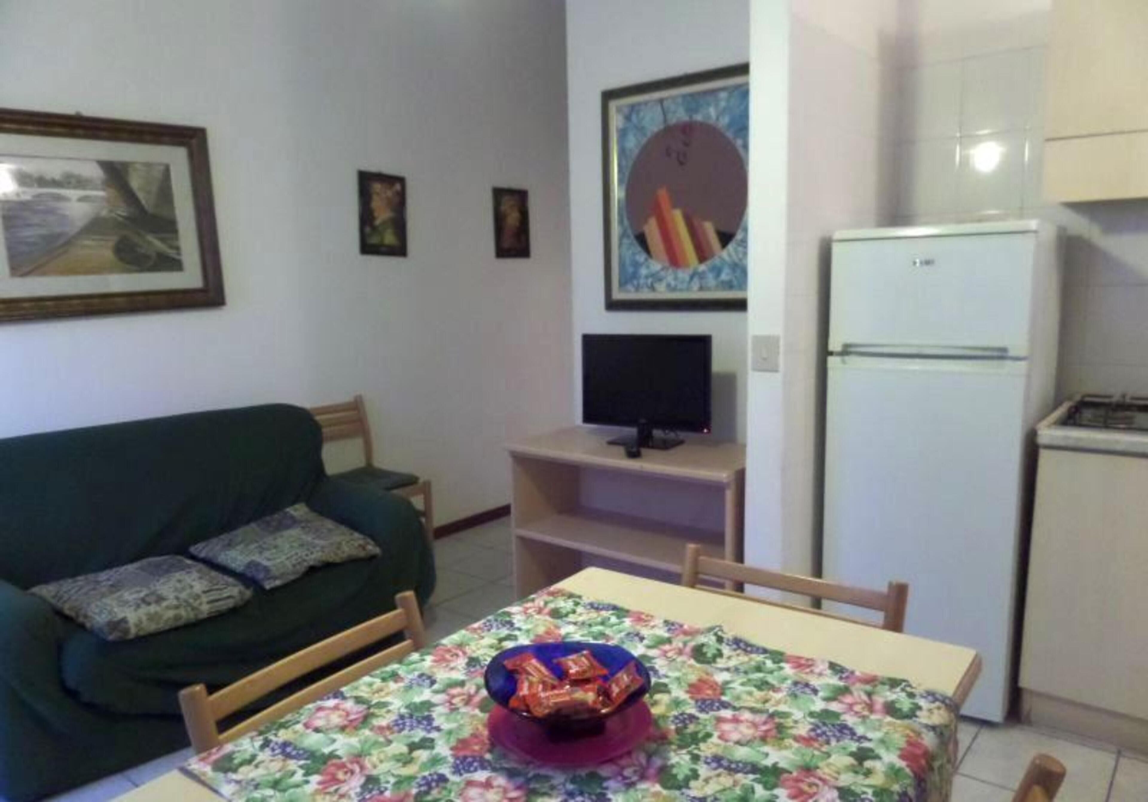 Un espai vital reduït amb un sofà verd, una taula de dinar amb estovell floral, un televisor sobre un suport i un frigorífic blanc.