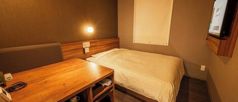 A compact hotel room with a single bed, a wooden desk with a telephone and a coffee maker, and a television mounted on the wall.