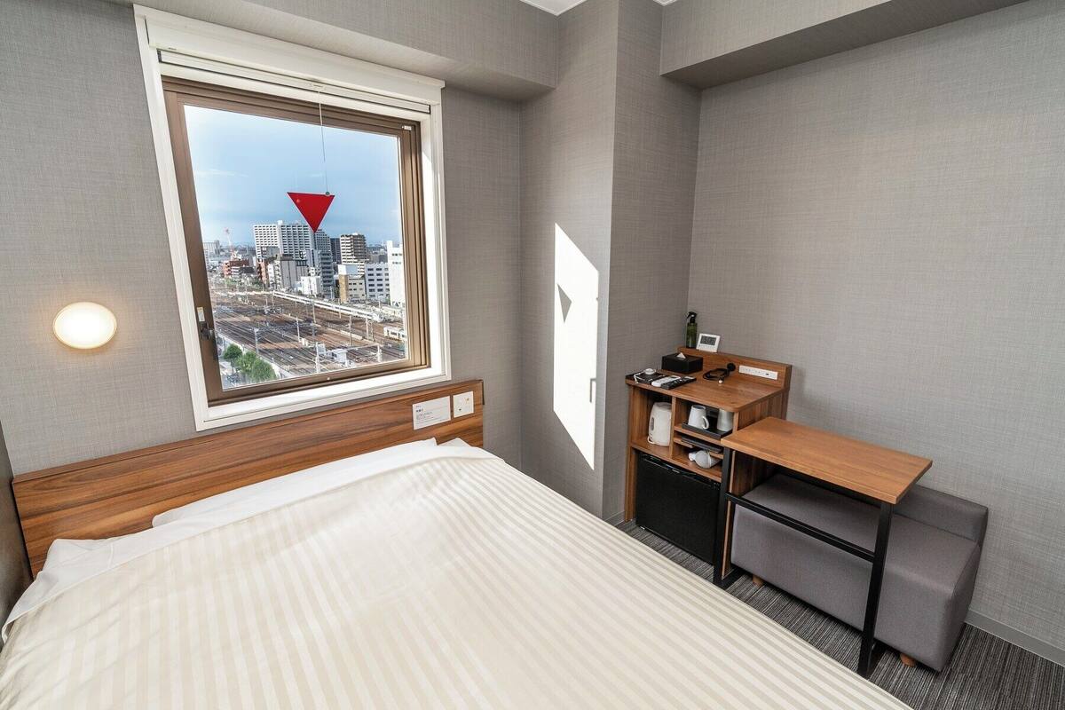 Habitación de hotel con cama, escritorio con silla, ventana con vista a la ciudad y lámpara fijada a la pared.