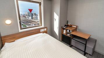 A hotel room with a bed, a desk with a chair, a window with a city view, and a wall-mounted light.