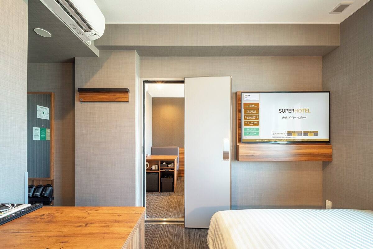 Habitación de hotel con cama, escritorio de madera, televisión montada en la pared y puerta que da a un baño y una kitchenette.