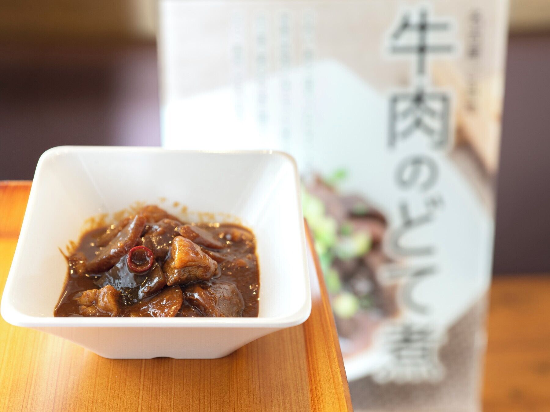 A white bowl of dark brown stew with visible pieces of meat and red chili peppers, placed on a wooden table.