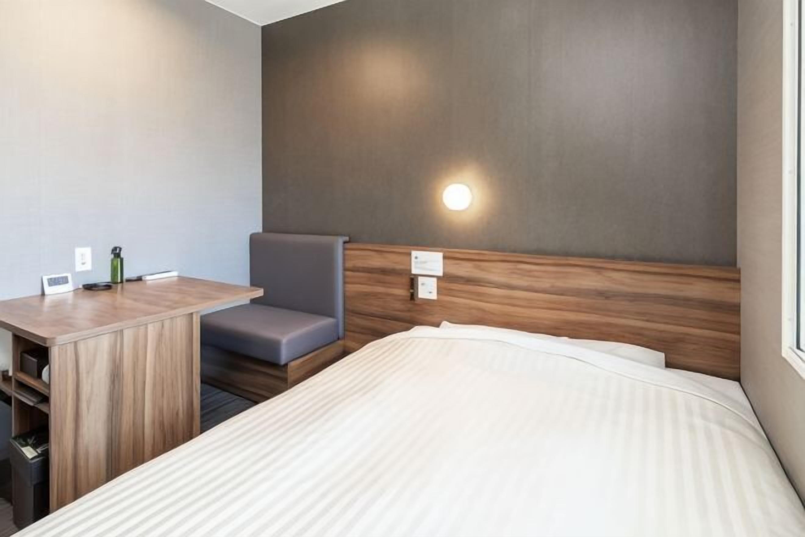 Et hotelværelse med seng, skrivebord, stol, natbord og vægmonteret lampe.