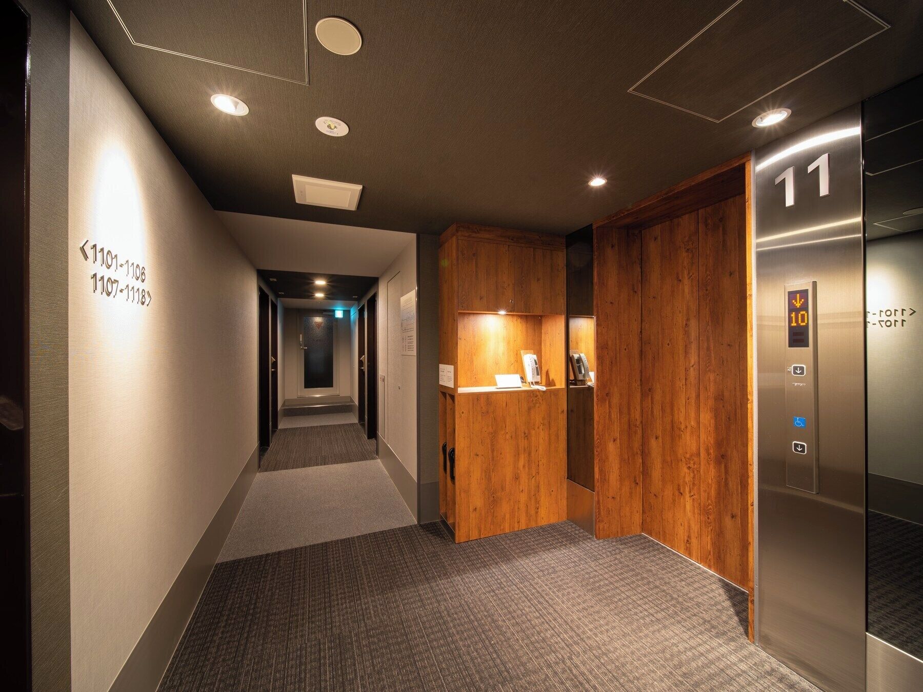 A modern hotel corridor with a wooden reception desk, an elevator, and directional signs.