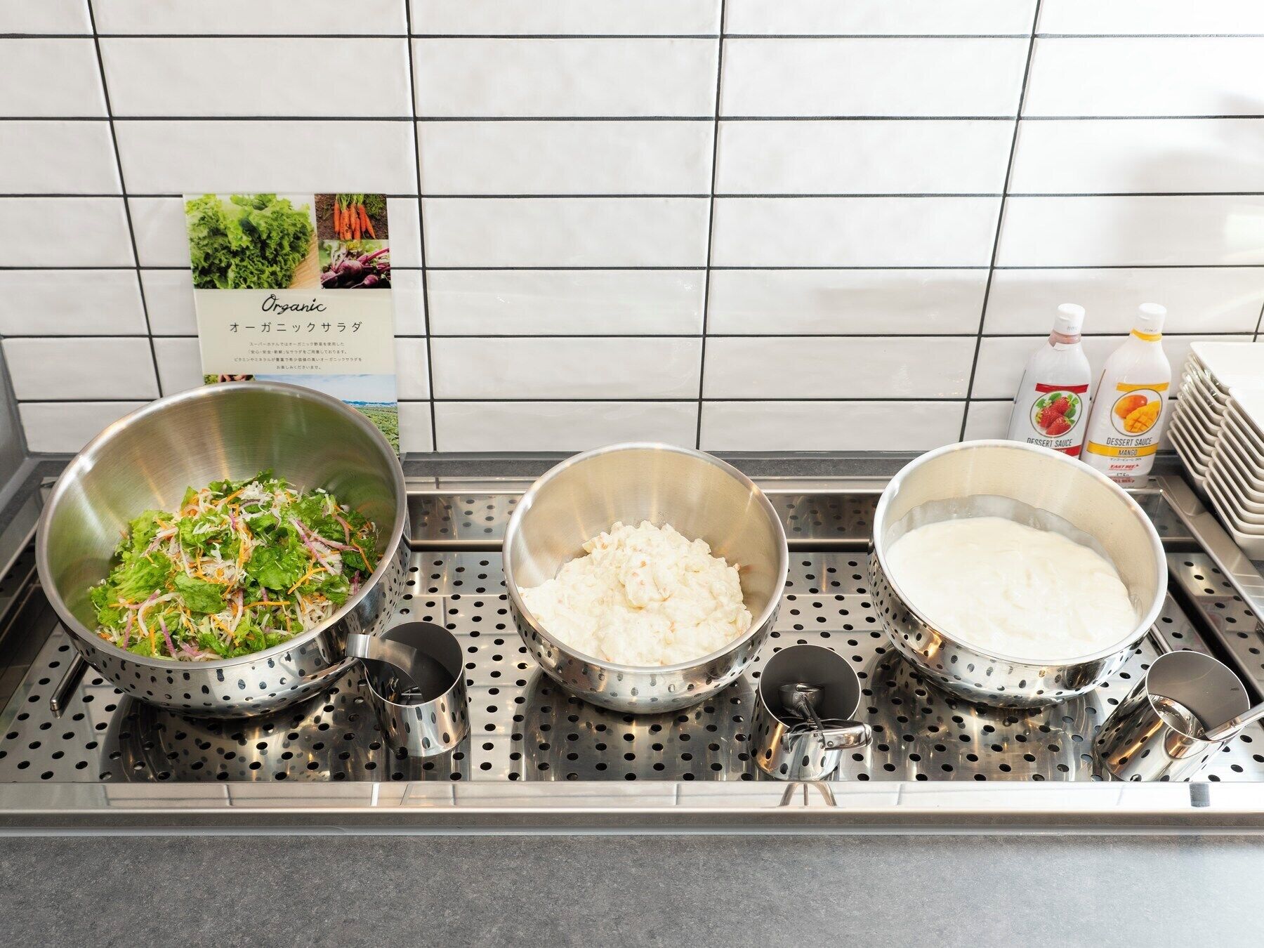Eine Küchenarbeitsplatte mit drei Edelstahl-Schüsseln, die verschiedene Lebensmittel, ein Sieb und zwei Flaschen Dessertsoße enthalten.