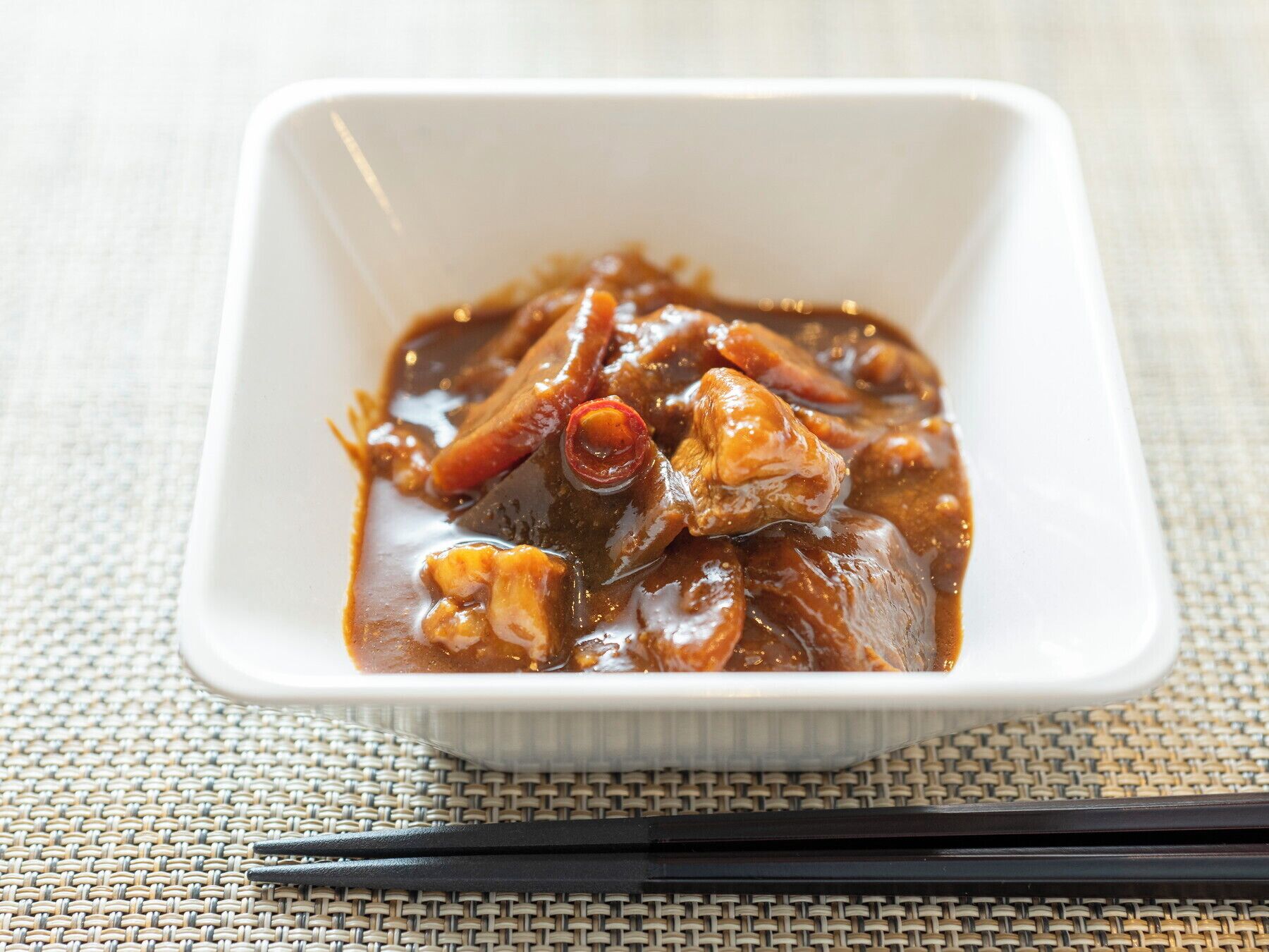 A white square dish containing a dark brown sauce with meat and vegetables, placed on a woven mat.