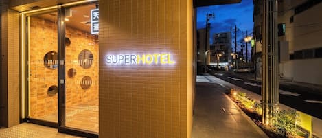 The exterior of a hotel with a neon sign reading "SUPERHOTEL" and a tiled entrance.