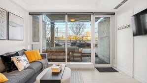A modern living room with a grey sofa, a coffee table, and a sliding glass door leading to an outdoor area with a bench and chairs. - Modern Ground-Level Suite UG Parking, AC, C-Train (Calgary)