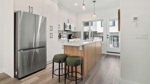 A modern kitchen with stainless steel appliances, a central island with stools, and a view to the living area. - Western Chic Retreat 3BR w/ 3 Ensuites, Prkg, AC (Calgary)