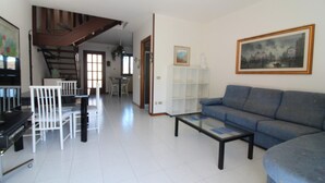 A spacious living room with a blue sofa, a dining area with white chairs, a staircase, and a painting on the wall.
